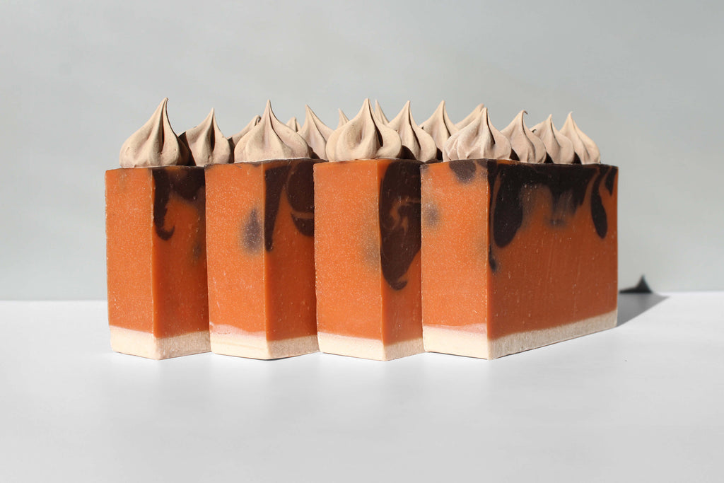Row of soaps with orange and brown swirl, and light brown frosting. On a light gray background.