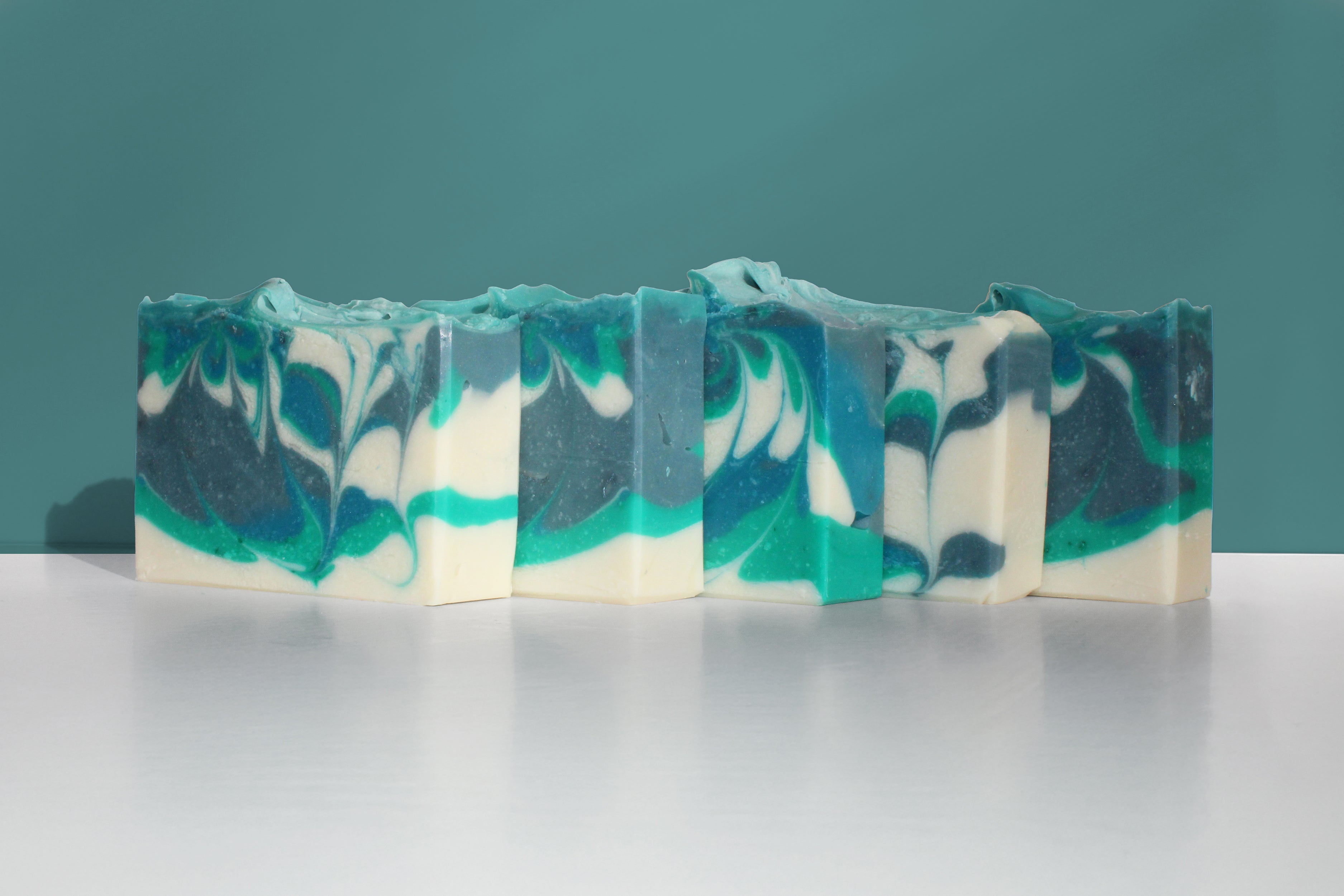 Four bars of swirled soap with green, white, and gray colors on a white surface against a teal background.